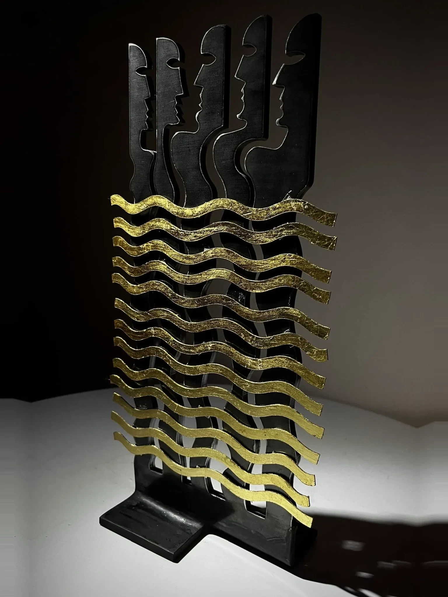 Flutti, sculpture de Gianluca Magnelli, cinq profils derrière des flots dorés.